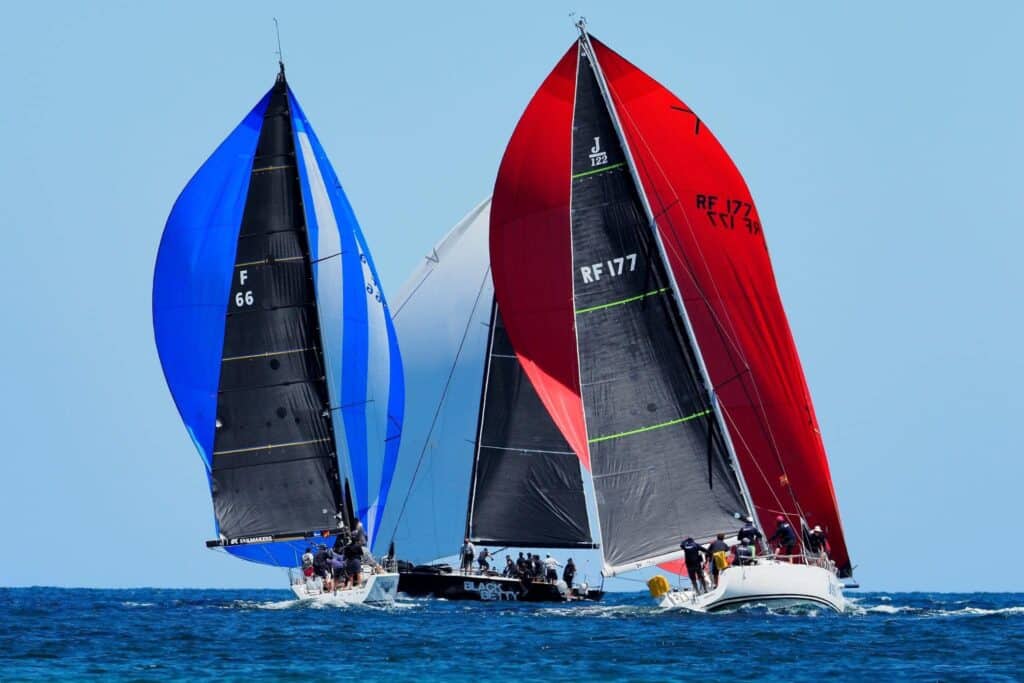 Checkmate and two other boats sailing downwind in close proximity with their spinnakers, blue, red and white.