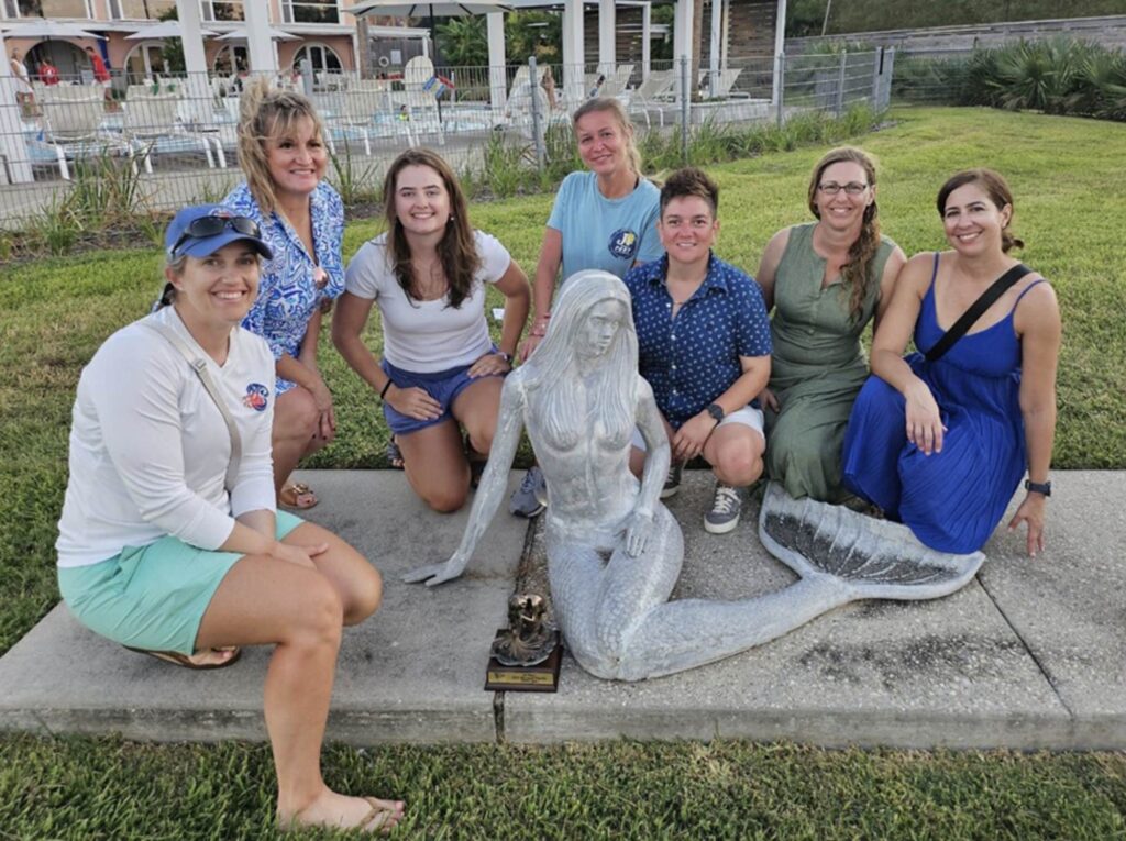 Meghan Pesch Mutlu and the BON BON crew pose with the Mermaid Perpetual Trophy and a mermaid statue at the Houston Yacht Club.